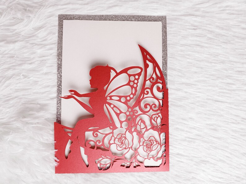 May include: Red laser cut invitation card with a fairy sitting on a crescent moon with roses and intricate designs. The card has a silver glitter border.