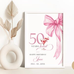 Può includere: Un cartello bianco stampabile con un fiocco rosa e un bicchiere da martini con una ciliegia. Il testo recita "50 anni amati, Buon compleanno Jane, 30 dicembre 20XX".