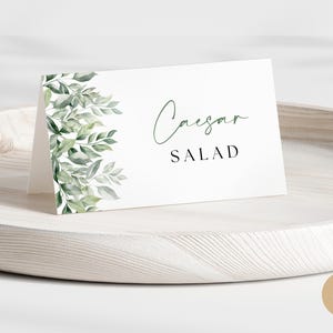 Pode incluir: Cartão branco com folhagem verde à esquerda e o texto "Salada Caesar" à direita.