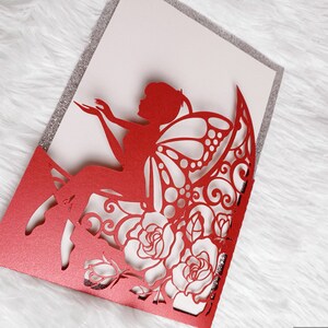 May include: Red laser cut invitation card with a fairy silhouette and floral design. The card is folded in half and has a glitter finish.