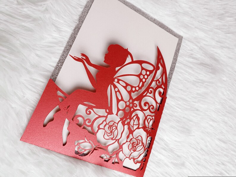 May include: Red laser cut invitation card with a fairy silhouette and floral design. The card is folded in half and has a glitter finish.
