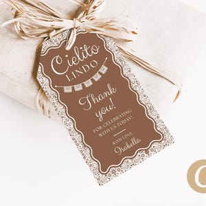 Peut inclure: Une étiquette cadeau marron avec une bordure blanche en dentelle et le texte "Cielito Lindo, Merci !" L'étiquette est attachée à un cadeau emballé avec un ruban de raphia. L'étiquette comprend également le nom "Isabella".
