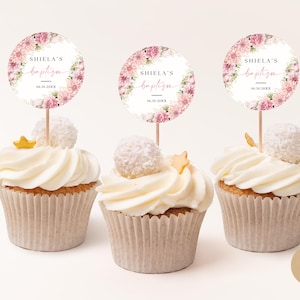 Pode incluir: Três cupcakes decorados com cobertura branca e granulado rosa e branco. Cada cupcake tem um topo redondo com padrão floral com o texto "Shiela's Baptism" e uma data.