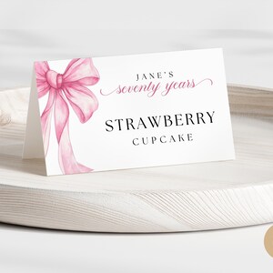 May include: A white place card with a pink bow and the text "Jane's seventy years Strawberry Cupcake".