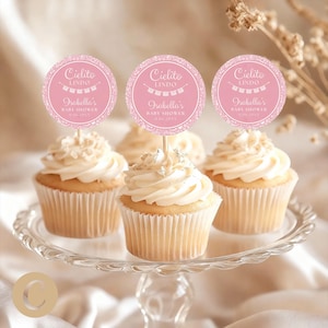 Peut inclure: Des cupcakes avec glaçage blanc et des décorations sur un présentoir à gâteau en verre. Les décorations sont roses avec le texte "Cielito Lindo" et "Isabella's Baby Shower". Les cupcakes sont dans des caissettes.