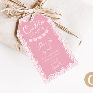 Peut inclure: Une étiquette cadeau rose avec une bordure en dentelle blanche et le texte "Cielito Lindo" et "Merci !" L'étiquette est attachée à un cadeau emballé avec un ruban de raphia. L'étiquette comprend également le nom "Isabella".