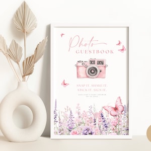 May include: A printable sign for a baby shower photo guestbook. The sign features a pink camera with a floral border and butterflies. The text reads "Photo Guestbook" and "Snap it. Shake it. Stick it. Sign it."