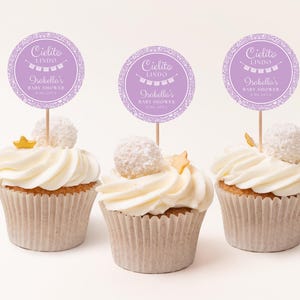Peut inclure: Trois cupcakes avec glaçage blanc, surmontés d'une boule de noix de coco et d'une étoile dorée. Chaque cupcake a un topper circulaire violet avec le texte "Cielito Lindo" et "Isabella's Baby Shower". Les cupcakes sont dans des caissettes en papier brun clair.