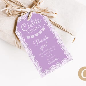 Peut inclure: Une étiquette cadeau lavande avec une bordure blanche en dentelle et le texte "Cielito Lindo, Merci !" attachée à un cadeau emballé avec du raphia. L'étiquette comprend le nom "Isabella".