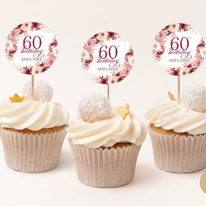 May include: Three cupcakes with white frosting and a white chocolate topping. Each cupcake has a round, pink and white floral themed topper with the text "60th birthday Adelaide".