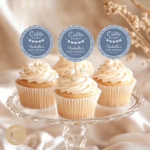 Peut inclure: Cupcakes avec glaçage blanc et décorations de baby shower. Les décorations sont rondes avec un fond bleu et le texte "Cielito Lindo" et "Isabella's Baby Shower". Les cupcakes sont présentés sur un présentoir à gâteau en verre.