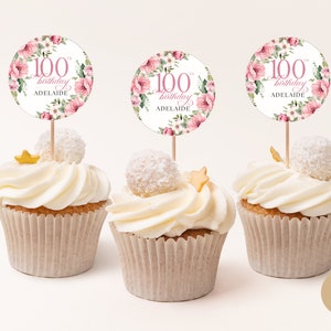 May include: Three cupcakes with white frosting and pink floral cupcake toppers that say "100th birthday Adelaide".