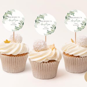 Pode incluir: Três cupcakes brancos com toppers florais verdes e brancos com o tema "Michael's baptism" com uma data. Os cupcakes são decorados com glacê branco e granulado dourado.