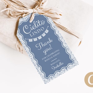 Peut inclure: Une étiquette cadeau bleue avec une bordure blanche ornée. Elle porte l'inscription "Cielito Lindo" et "Thank you!" en écriture cursive. Le cadeau est emballé dans du papier brun et noué avec du raphia.