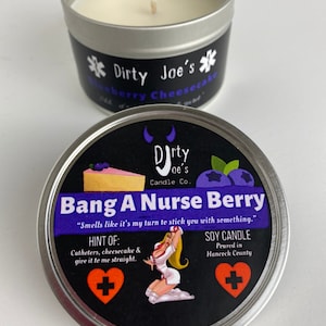 Könnte beinhalten: Eine silberne Blechdose mit einer schwarzen Etikette, auf der "Dirty Joe's Candle Co. Bang A Nurse Berry" und "Smells like it's my turn to stick you with something." steht. Das Etikett enthält auch eine Zeichnung einer Krankenschwester in weißer Uniform und ein rotes Herz mit einem weißen Pluszeichen. Die Kerze ist eine Sojawachskerze, die im Hanceck County gegossen wurde.