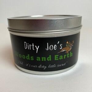 May include: A round, silver-lidded tin with a black label. The label reads "Dirty Joe's Woods and Earth" in white and green text, with a deer graphic. The phrase "Shh... it's our dirty, little secret" is also printed.