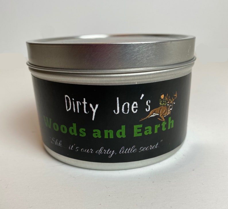May include: A round, silver-lidded tin with a black label. The label reads "Dirty Joe's Woods and Earth" in white and green text, with a deer graphic. The phrase "Shh... it's our dirty, little secret" is also printed.