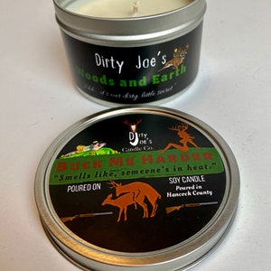 May include: A soy candle in a silver tin with a black label. The label reads "Dirty Joe's Woods and Earth" and "Buck Me Harder." The lid features a deer graphic and the text "Smells like, someone's in heat."