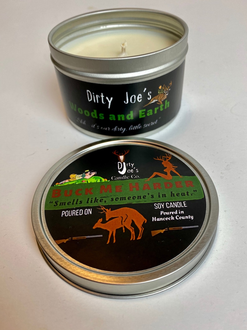 May include: A soy candle in a silver tin with a black label. The label reads "Dirty Joe's Woods and Earth" and "Buck Me Harder." The lid features a deer graphic and the text "Smells like, someone's in heat."