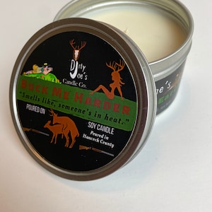 May include: A soy candle in a silver tin with a black label. The label reads "BUCK ME HARDER" with deer graphics and the text "Smells like, someone's in heat." The candle is from Dirty Joe's Candle Co.
