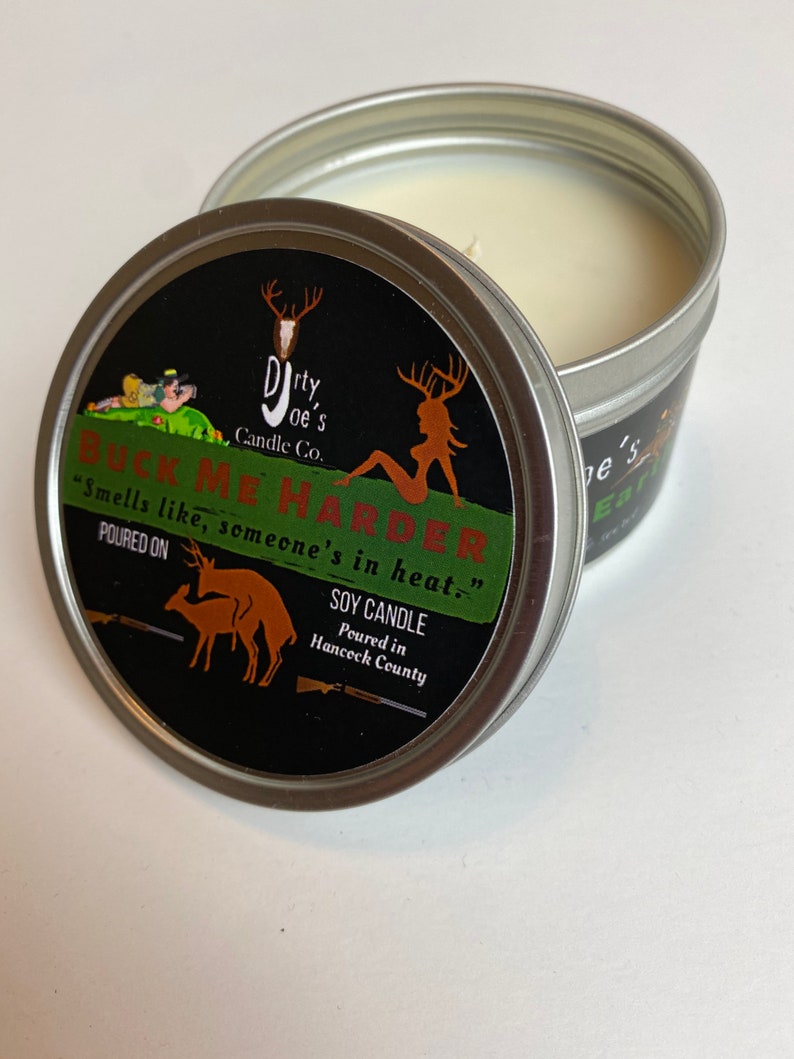 May include: A soy candle in a silver tin with a black label. The label reads "BUCK ME HARDER" with deer graphics and the text "Smells like, someone's in heat." The candle is from Dirty Joe's Candle Co.