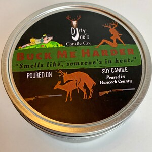May include: A round, silver-colored candle tin with a black label. The label features the text "BUCK ME HARDER" and "Smells like, someone's in heat." along with deer illustrations and the words "SOY CANDLE". The candle is from Dirty Joe's Candle Co.