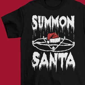 May include: Black t-shirt with the text "SUMMON SANTA" in a distressed white font. The design includes a pentagram with a Santa hat in the center. The shirt is a crew neck and has short sleeves.