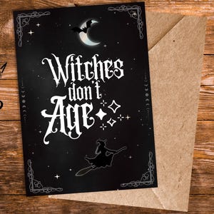 Pode incluir: Um cartão preto com o texto branco "Witches don't Age" em letras góticas, uma lua crescente e uma bruxa numa vassoura. O cartão está colocado num envelope castanho, sobre um fundo de madeira.