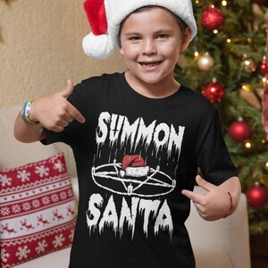 May include: A young person wearing a black t-shirt with the words "SUMMON SANTA" in a gothic font, along with a pentagram and a Santa hat graphic. The person is also wearing a red and white Santa hat.