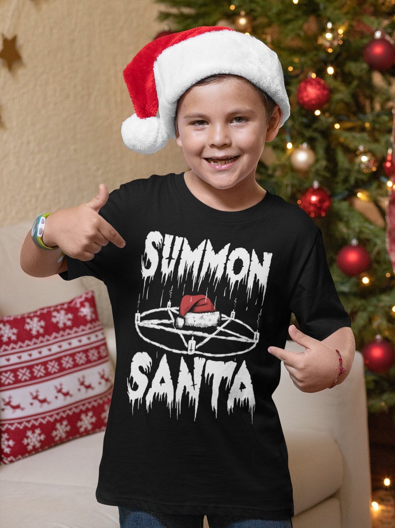 May include: A young person wearing a black t-shirt with the words "SUMMON SANTA" in a gothic font, along with a pentagram and a Santa hat graphic. The person is also wearing a red and white Santa hat.
