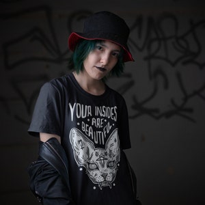 May include: A person with green hair wearing a black t-shirt with a white graphic of a cat with the text "Your insides are beautiful". The person is also wearing a black hat with a red brim and a black leather jacket.