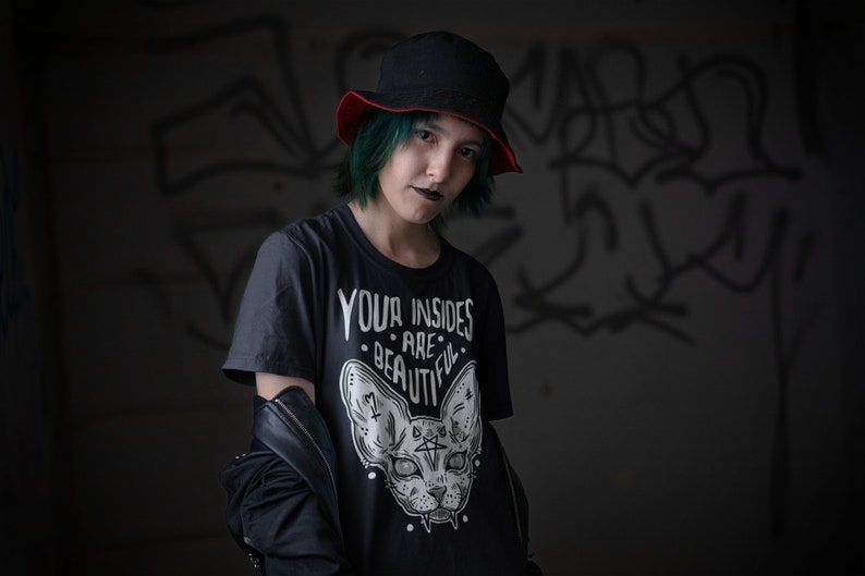 May include: A person with green hair wearing a black t-shirt with a white graphic of a cat with the text "Your insides are beautiful". The person is also wearing a black hat with a red brim and a black leather jacket.