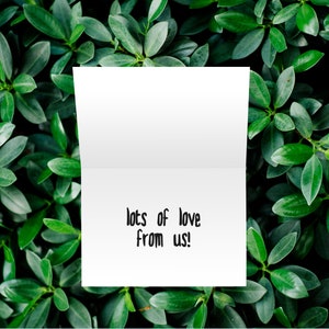 May include: A white card with the text "lots of love from us!" written in black ink. The card is lying on a bed of green leaves.
