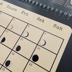 May include: A close-up of a spiral-bound calendar with a tan background and black grid lines. The calendar displays the days of the week, with the phases of the moon illustrated for each date. The text "Thurs", "Fri", "Sat", and "Sun" are visible.
