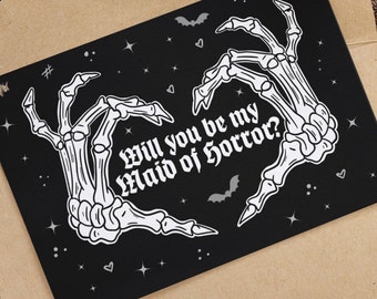 May include: A black card with a white skeleton hand design forming a heart shape. The text "Will you be my Maid of Horror?" is written inside the heart.