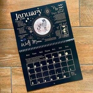 May include: A black 2026 Wolf Moon calendar with a detailed illustration of a wolf inside a circle. The calendar includes a monthly calendar grid, lunar phases, and text about energies, spell work, and power crystals.