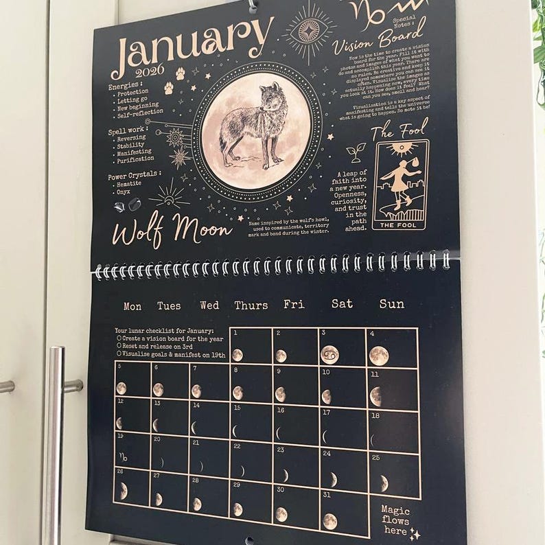 May include: Black and gold wall calendar for January 2020, featuring a wolf illustration and moon phase details. Includes text: "Wolf Moon", "Vision Board", and "The Fool". The calendar has a spiral binding and is hanging on a white door.