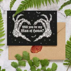 May include: A black card with a white skeleton hand design forming a heart shape. The text reads "Will you be my Man of Honor?" with a black background and white stars.