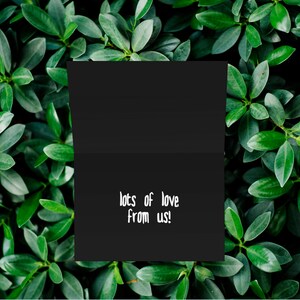 May include: A black card with the text "lots of love from us!" written in white on it. The card is surrounded by green leaves.