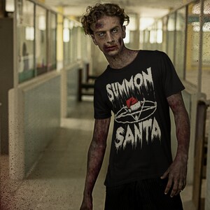 May include: A black t-shirt with the text "SUMMON SANTA" in a distressed, gothic font, featuring a pentagram and Santa hat design. The image has a horror theme, with a zombie-like figure in a hallway.