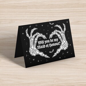 May include: Black card with white skeleton hands forming a heart shape. The text reads "Will you be my Maid of Honour?" with stars and bats in the background.