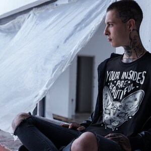 May include: A young person wearing a black t-shirt with a white graphic of a cat's face and the text "YOUR INSIDES ARE BEAUTIFUL". The person is sitting on a ledge with a white plastic sheet behind them.