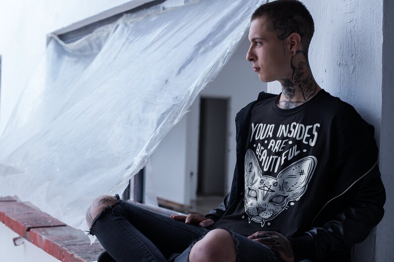 May include: A young person wearing a black t-shirt with a white graphic of a cat's face and the text "YOUR INSIDES ARE BEAUTIFUL". The person is sitting on a ledge with a white plastic sheet behind them.