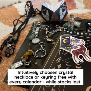 May include: An assortment of crystal keyrings and necklaces, alongside a small booklet and stickers. The keyrings feature various colored stones and charms. The stickers depict celestial and mystical imagery. The text on the image reads: "Intuitively chosen crystal necklace or keyring free with every calendar - while stocks last."