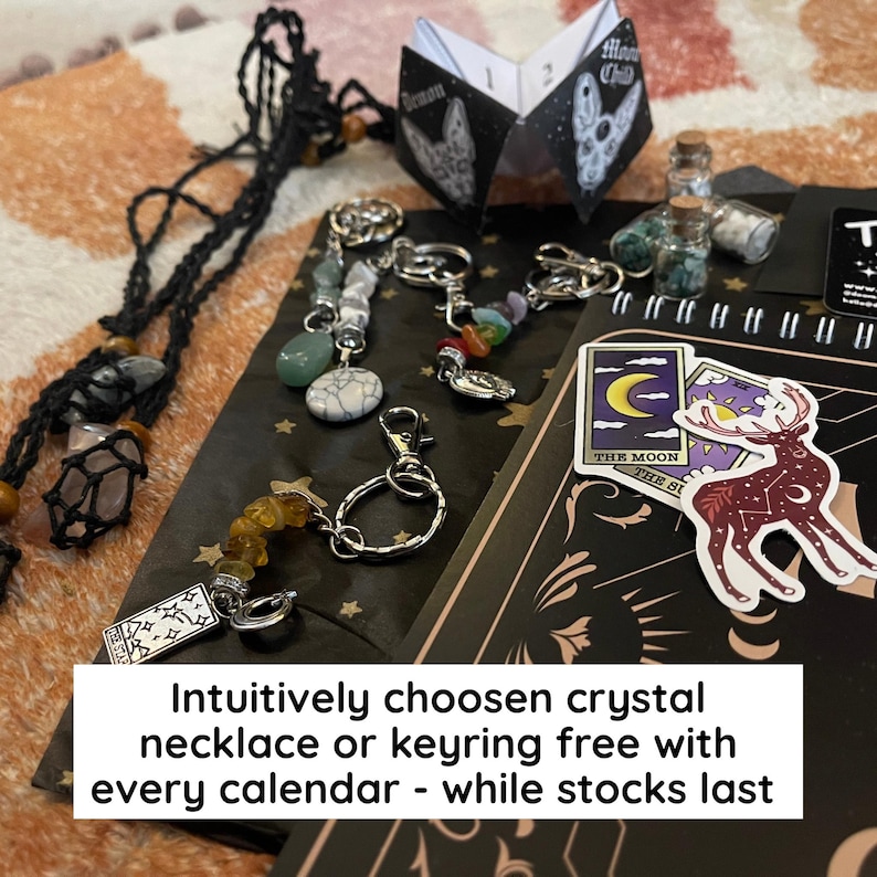 May include: An assortment of crystal keyrings and necklaces, alongside a small booklet and stickers. The keyrings feature various colored stones and charms. The stickers depict celestial and mystical imagery. The text on the image reads: "Intuitively chosen crystal necklace or keyring free with every calendar - while stocks last."