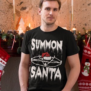 May include: Black t-shirt with the text "SUMMON SANTA" in a dripping font, featuring a pentagram with a Santa hat in the center. The shirt is worn by a person in front of a fireplace decorated for Christmas.
