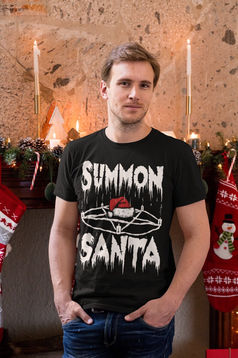 May include: Black t-shirt with the text "SUMMON SANTA" in a dripping font, featuring a pentagram with a Santa hat in the center. The shirt is worn by a person in front of a fireplace decorated for Christmas.