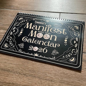 May include: A black and white spiral-bound calendar titled "Doomcat's Manifest with the Moon Calendar 2026." The calendar features celestial and geometric designs. The cover is black with white lettering and illustrations.