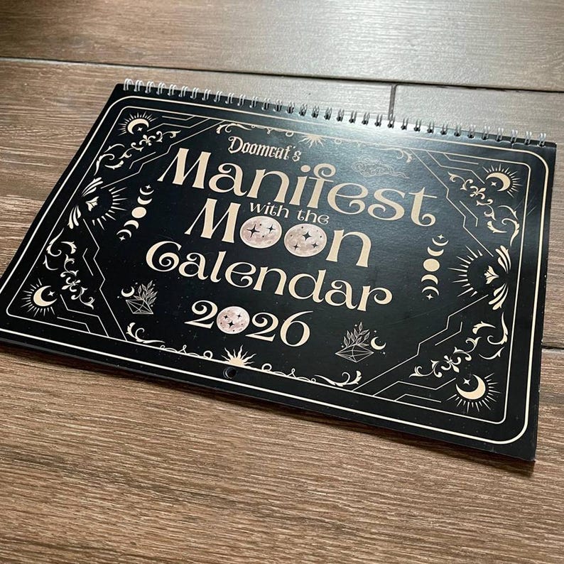 May include: A black and white spiral-bound calendar titled "Doomcat's Manifest with the Moon Calendar 2026." The calendar features celestial and geometric designs. The cover is black with white lettering and illustrations.
