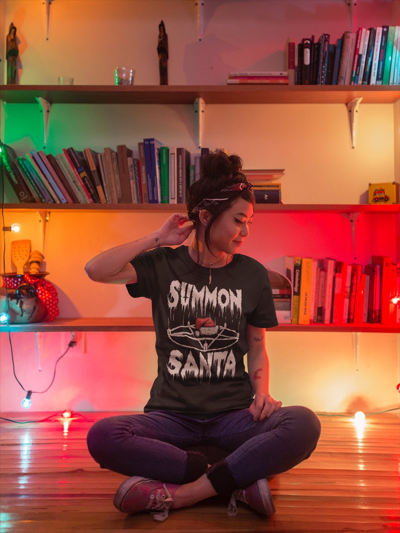 May include: A dark green t-shirt with the text "SUMMON SANTA" in a dripping, gothic font, featuring a pentagram and Santa hat design. The person is wearing blue jeans and pink sneakers, sitting on a wooden floor with bookshelves in the background.